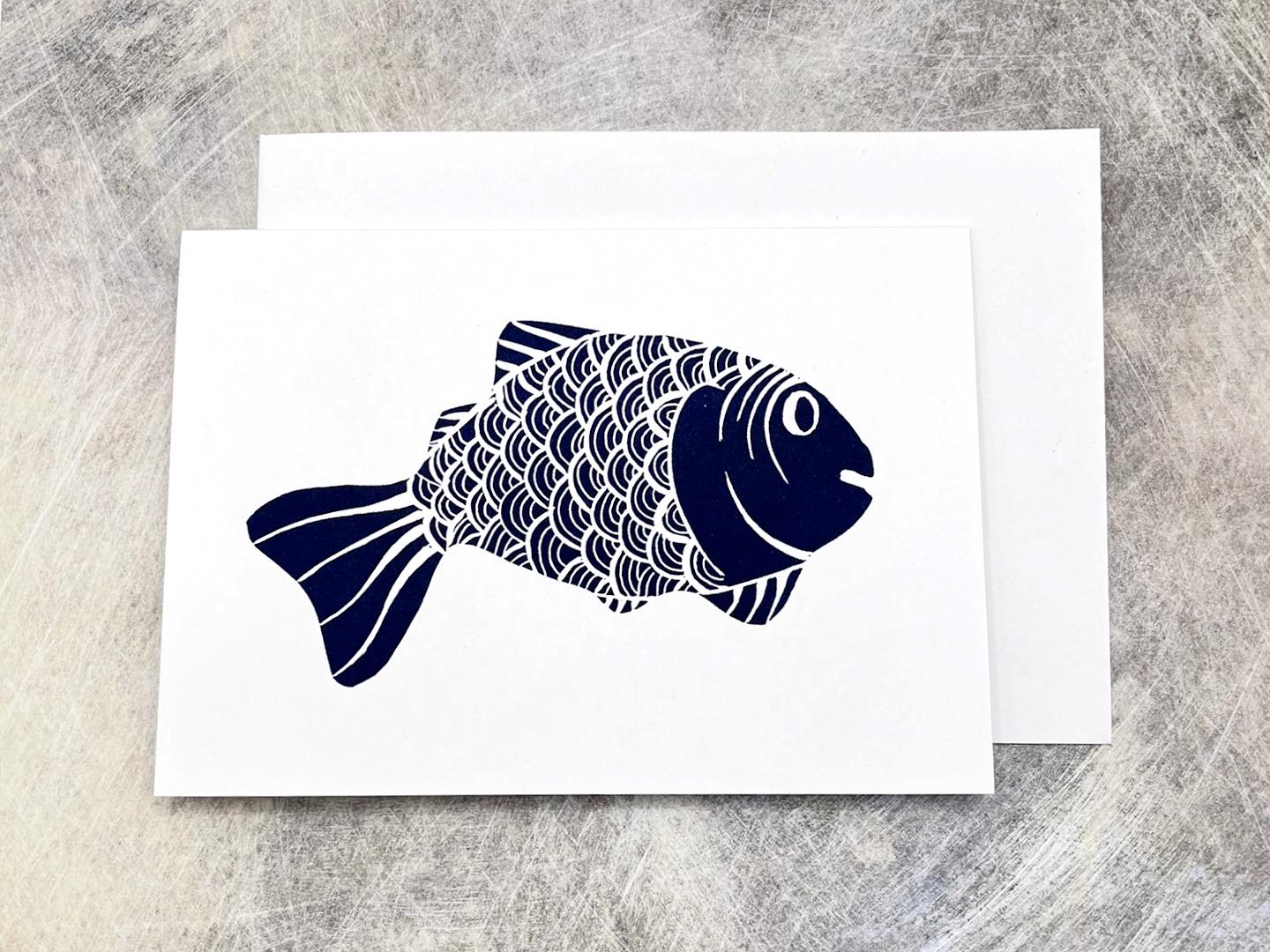 White card with an original lino printed blue fish, with matching envelope, laid on a textured background. 