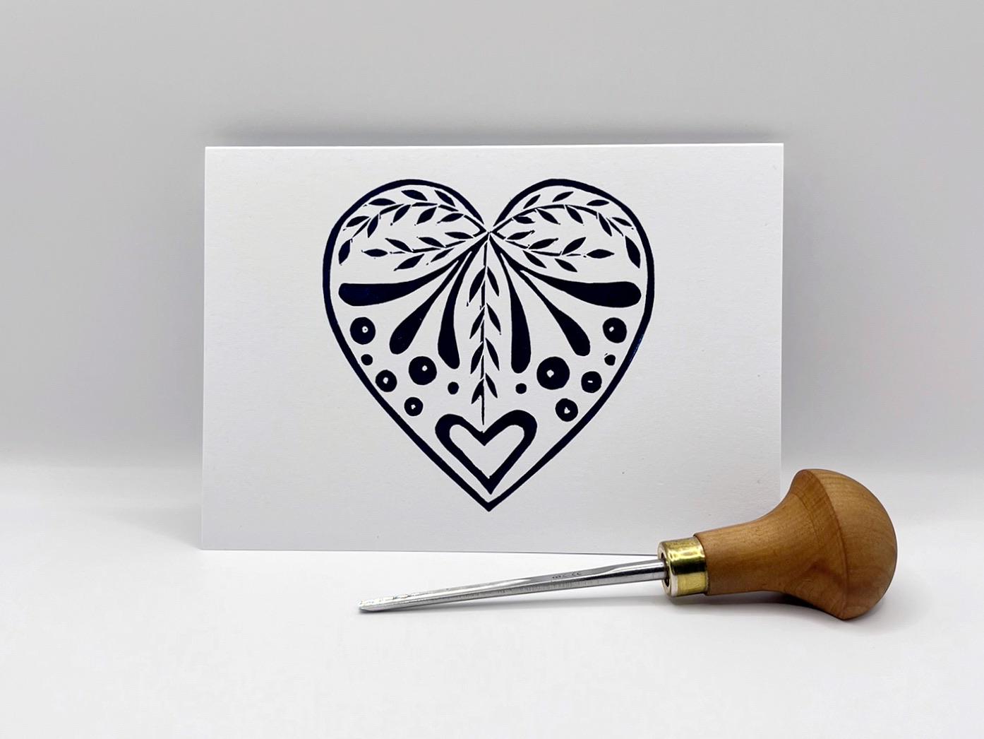 White card with an original lino printed  blue heart design stood on a white surface. Photo includes lino cutting tool.