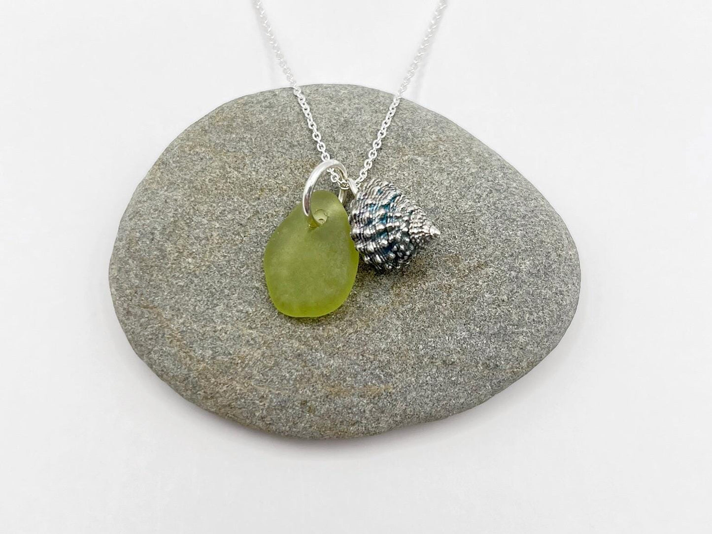 Necklace with a lime green sea glass and a silver shell charm laid on a grey pebble