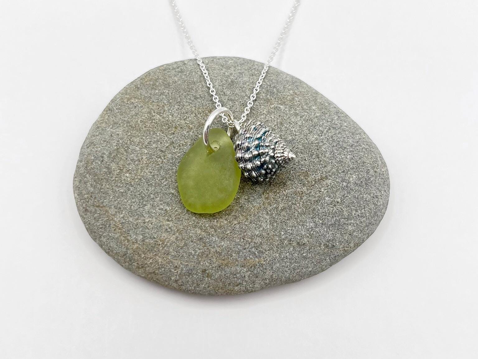 Necklace with a lime green sea glass and a silver shell charm laid on a grey pebble