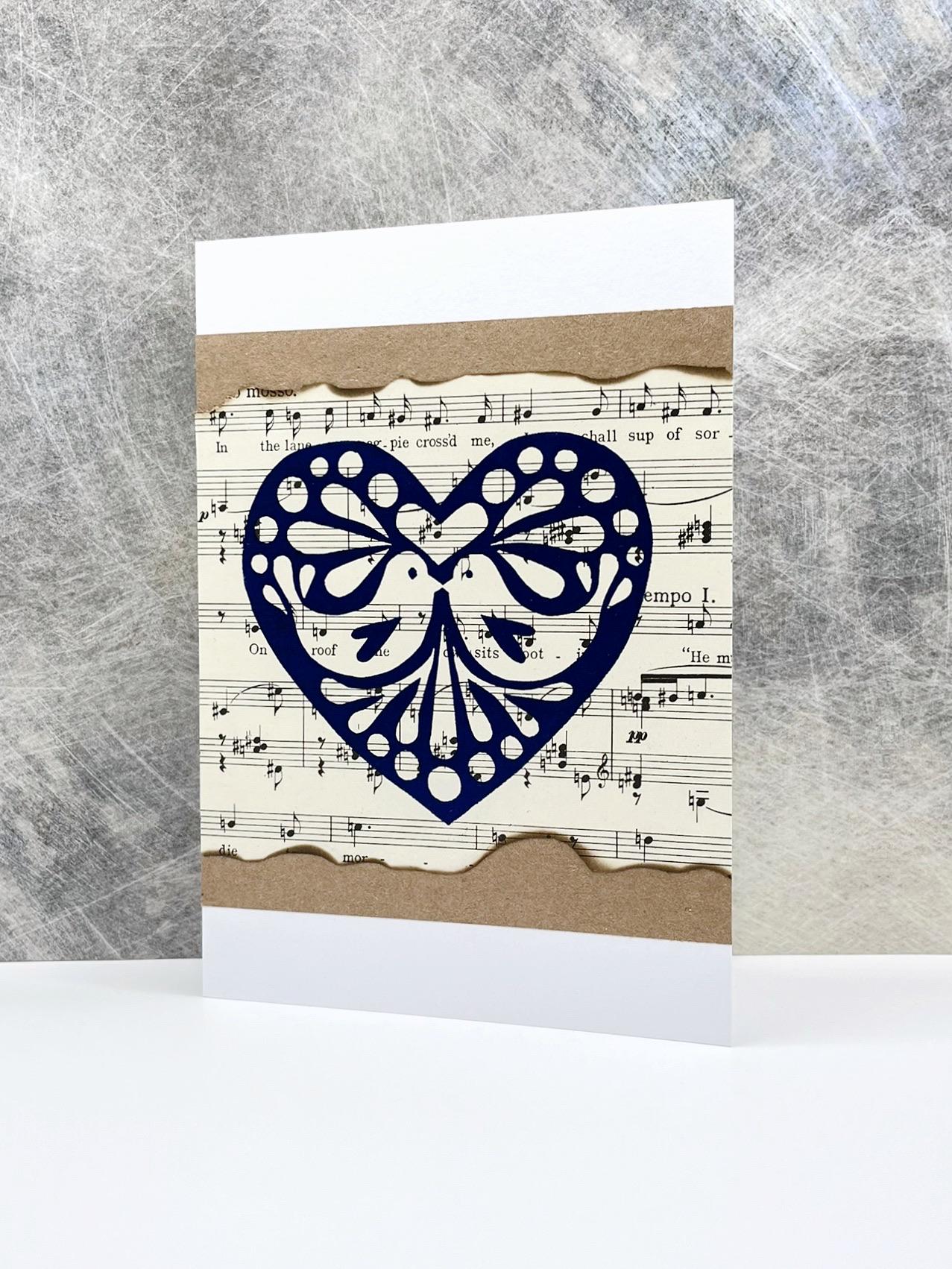 Greeting card with an original lino printed blue heart design on sheet music, stood against a textured background.