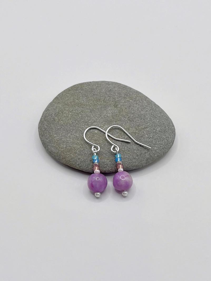Pair of pastel seed bead and lilac jasper earrings laid on a grey pebble against a white background.