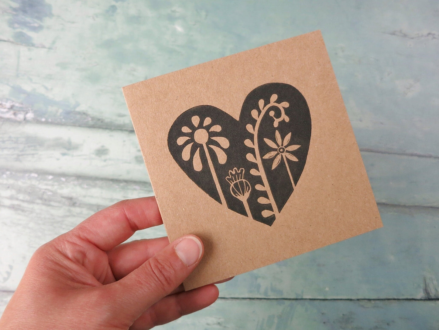 Kraft paper card with an original lino printed floral heart design held in a hand against a textured surface. 