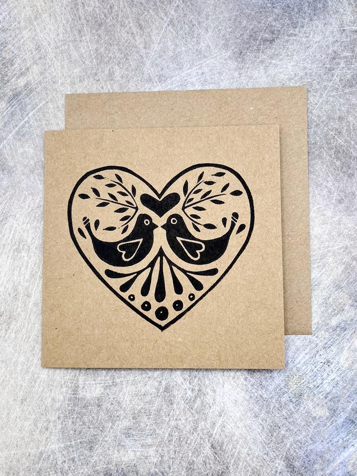 Kraft card with black lino print heart design featuring two birds laid on a textured grey background