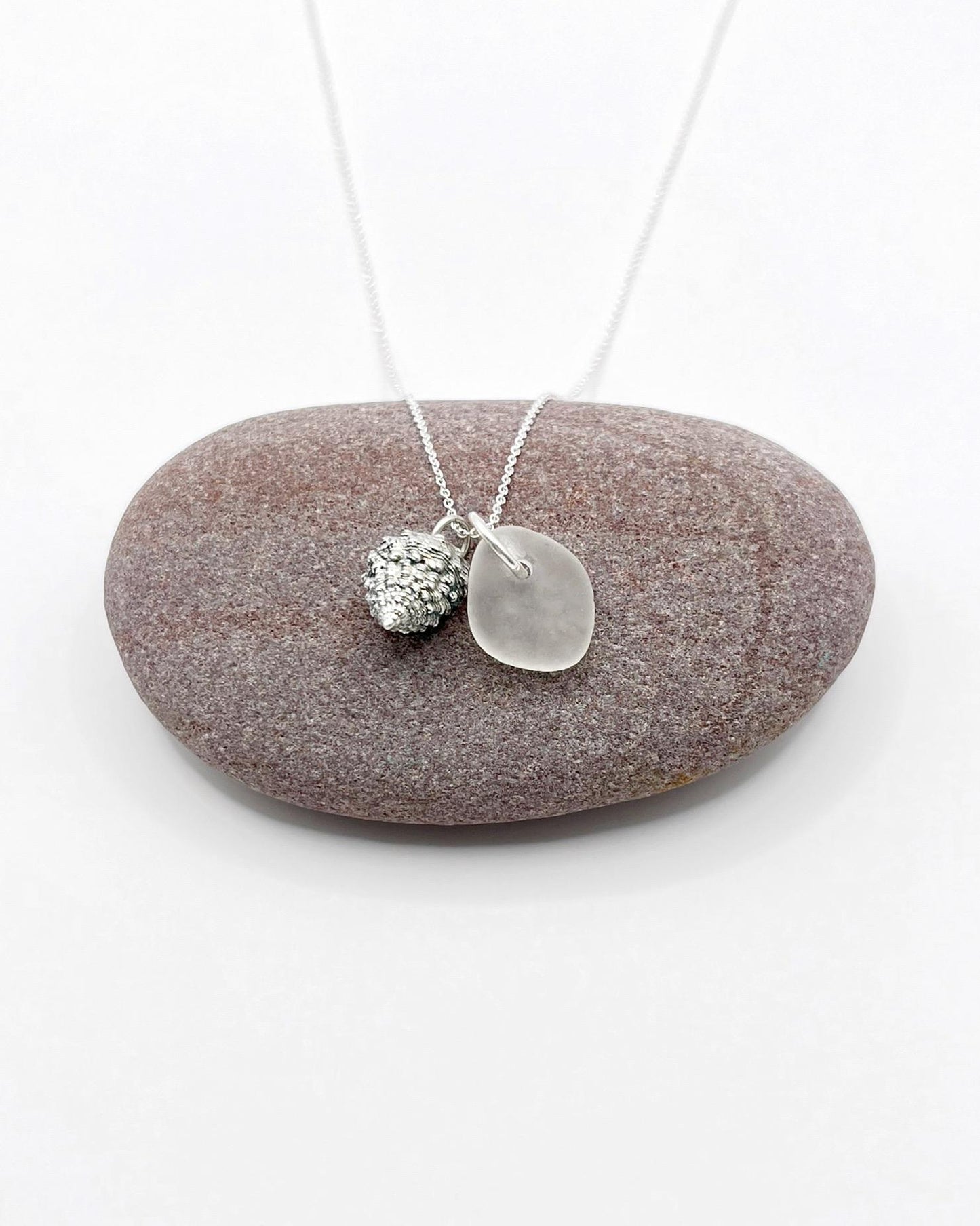 Silver necklace with White Sea glass and silver shell pendant laid on a purple pebble against a white background