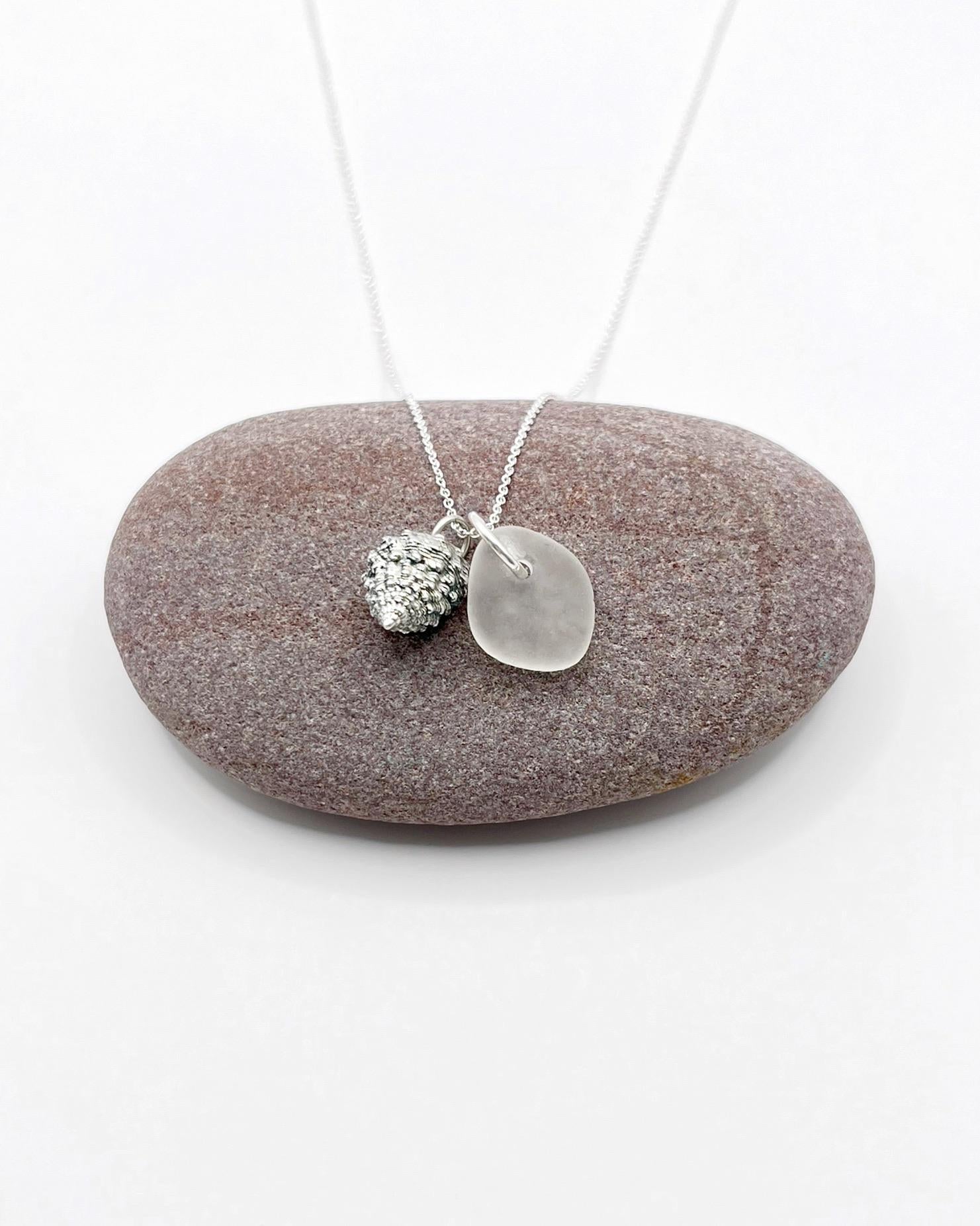Silver necklace with White Sea glass and silver shell pendant laid on a purple pebble against a white background