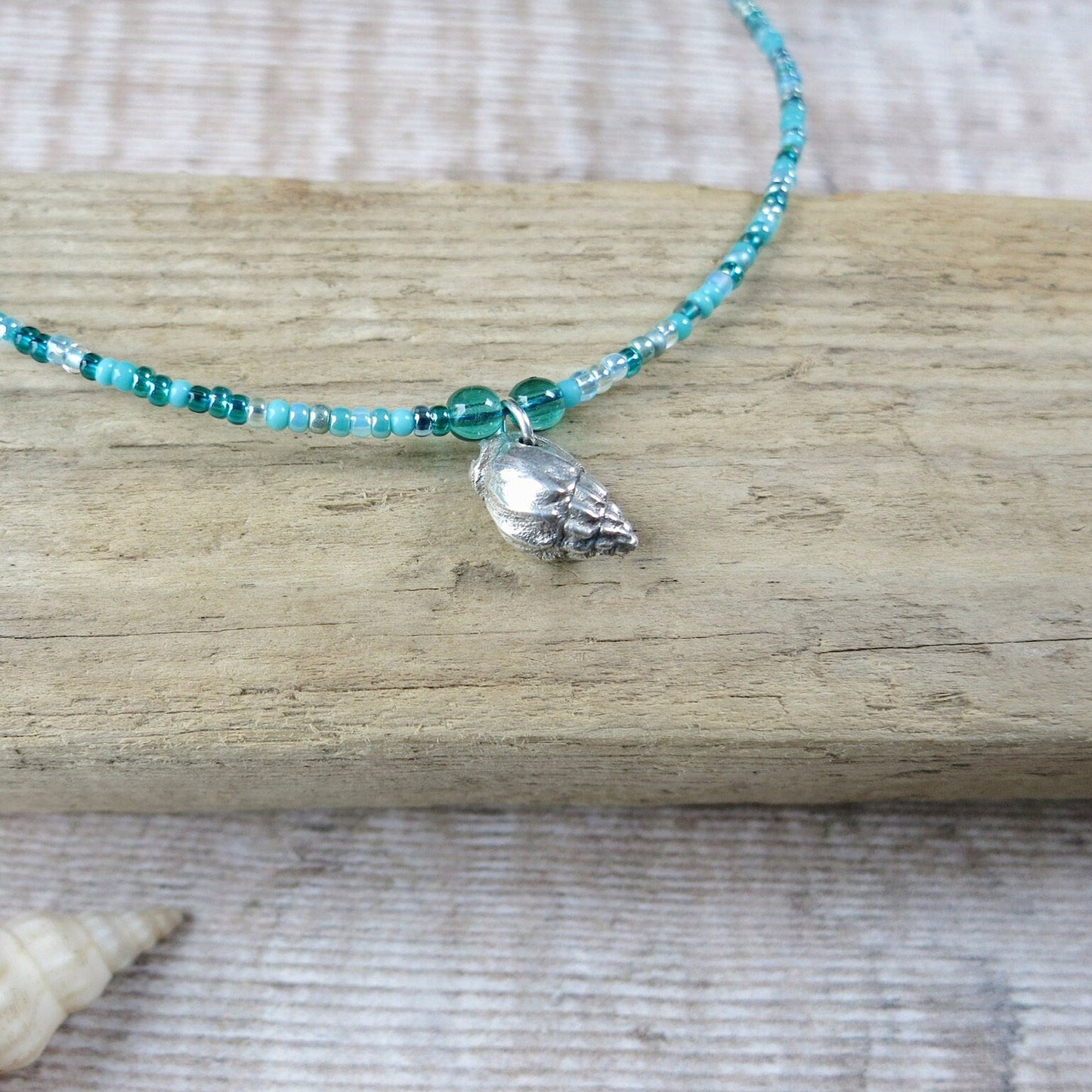 Turquoise seed bead necklace with a silver shell pendant laid on driftwood against a textured background