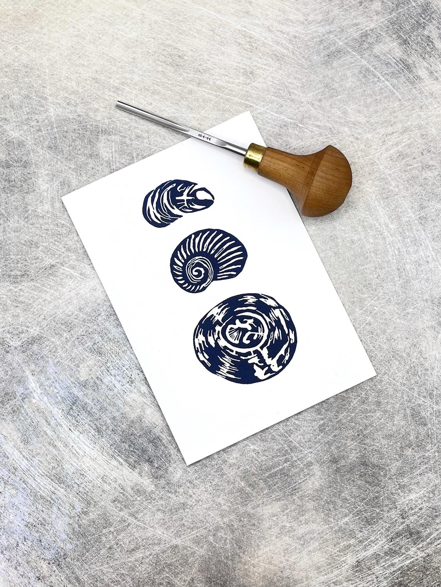 Three blue original lino printed shells on white card laid on a grey textured surface. Included in the photo is the lino cutting tool used.
