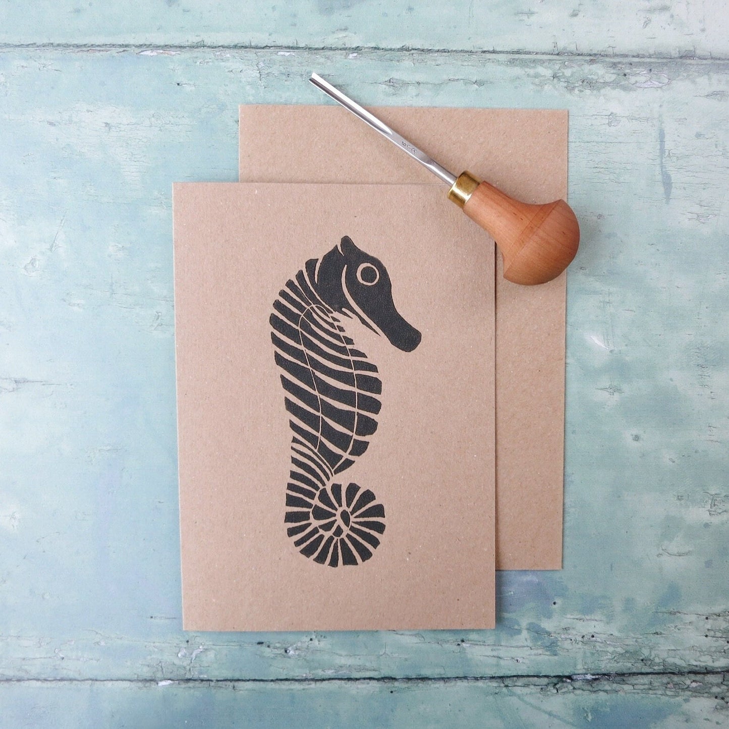 Kraft paper card with an original seahorse lino print laid on a textured blue surface. Photo includes lino cutting tool