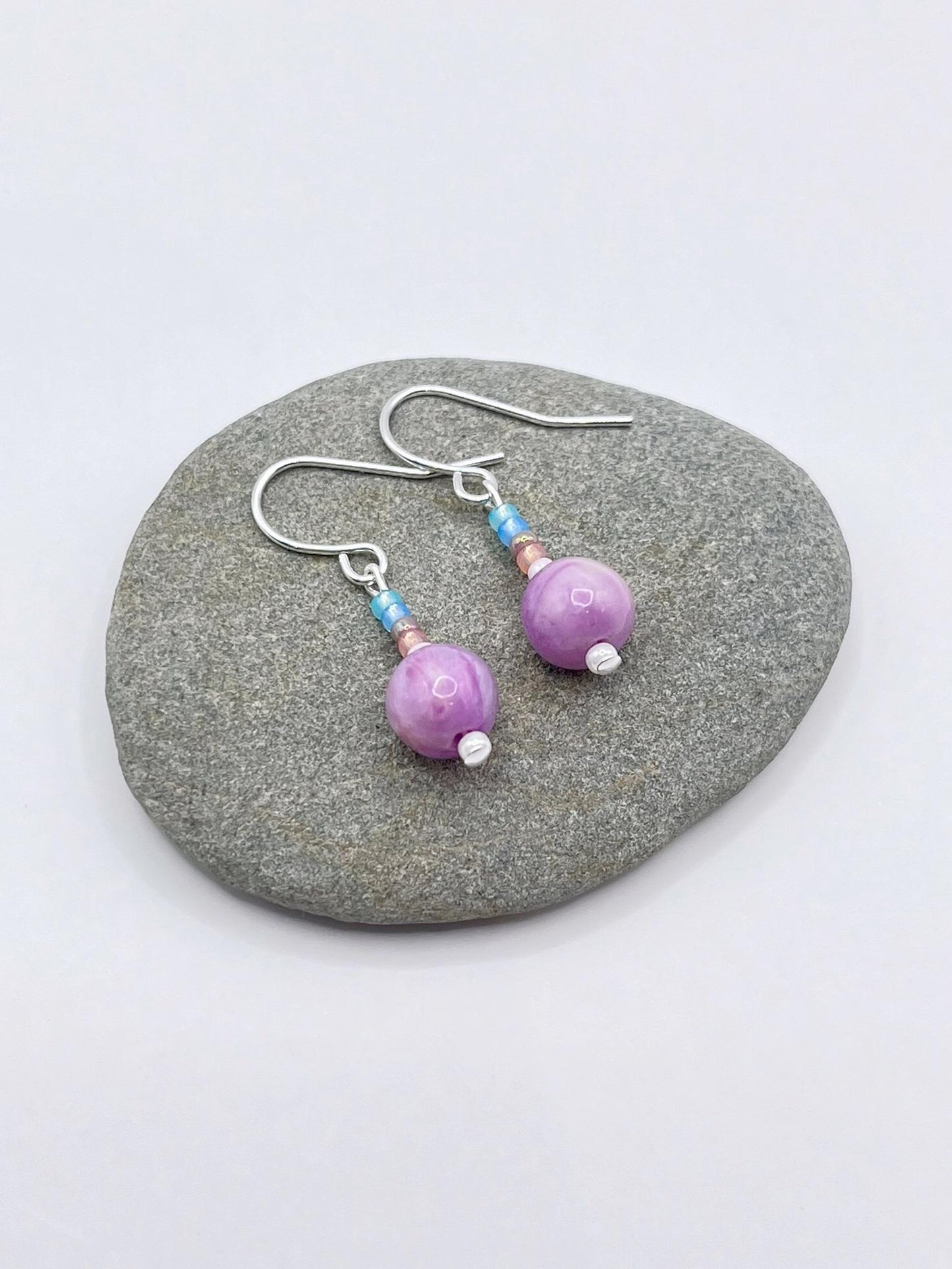 Pair of pastel seed bead and lilac jasper earrings laid on a grey pebble against a white background.