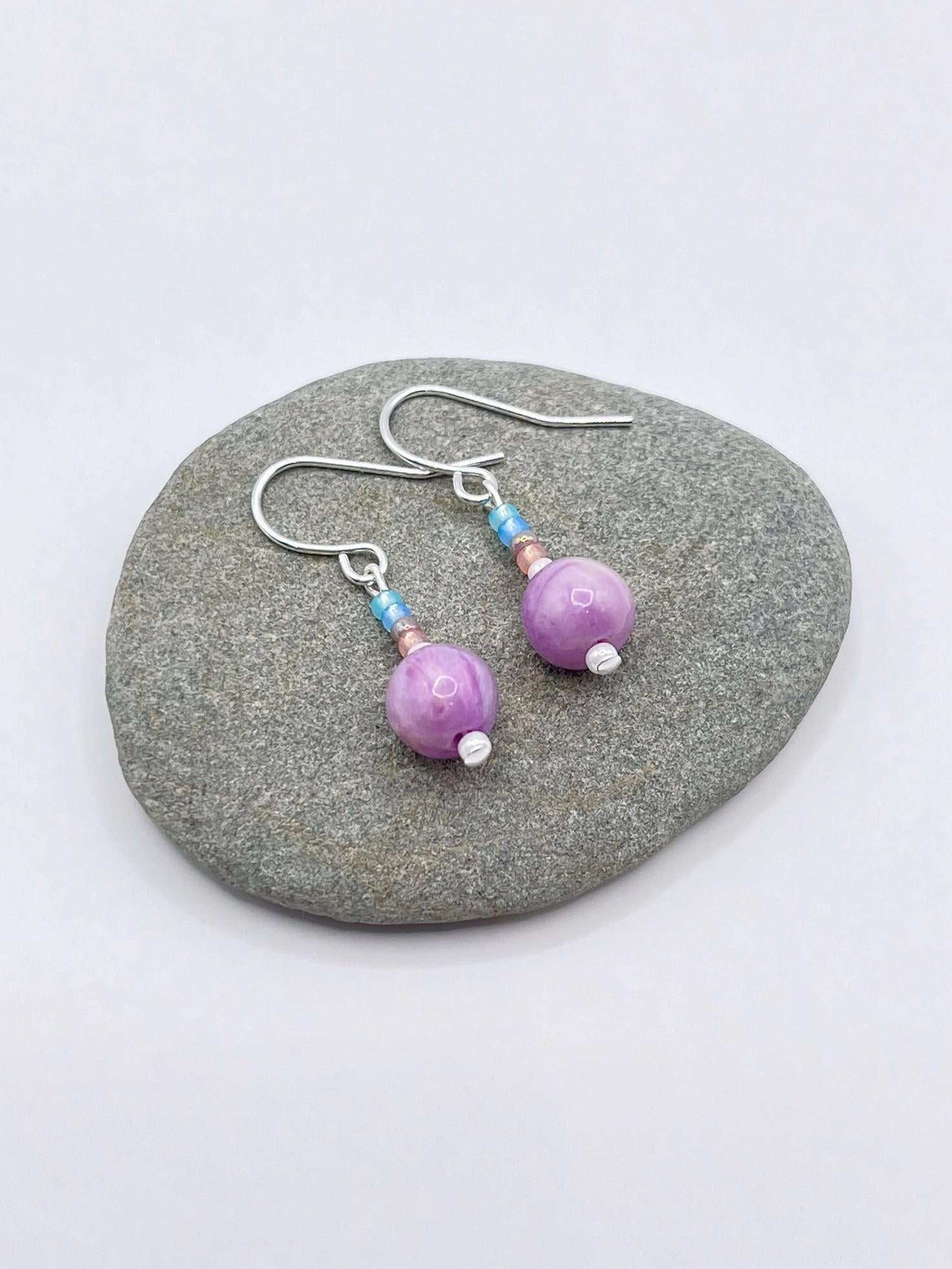 Pair of pastel seed bead and lilac jasper earrings laid on a grey pebble against a white background.
