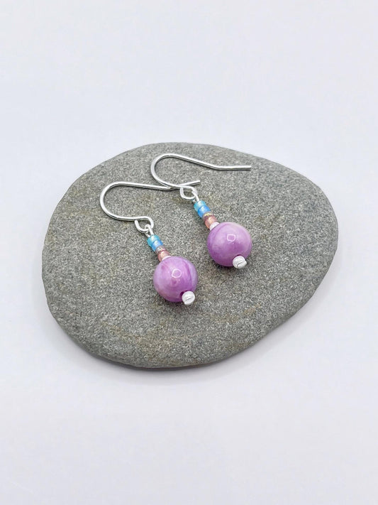 Pair of pastel seed bead and lilac jasper earrings laid on a grey pebble against a white background.