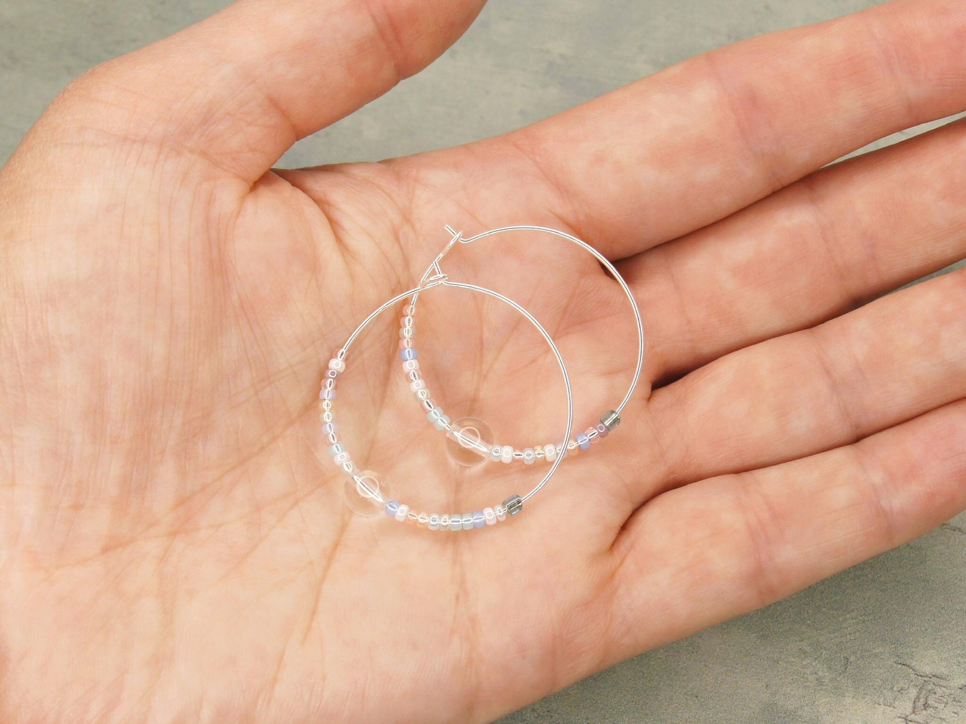 Pair of pastel seed bead and clear crystal hoop earrings held in a hand against a green background