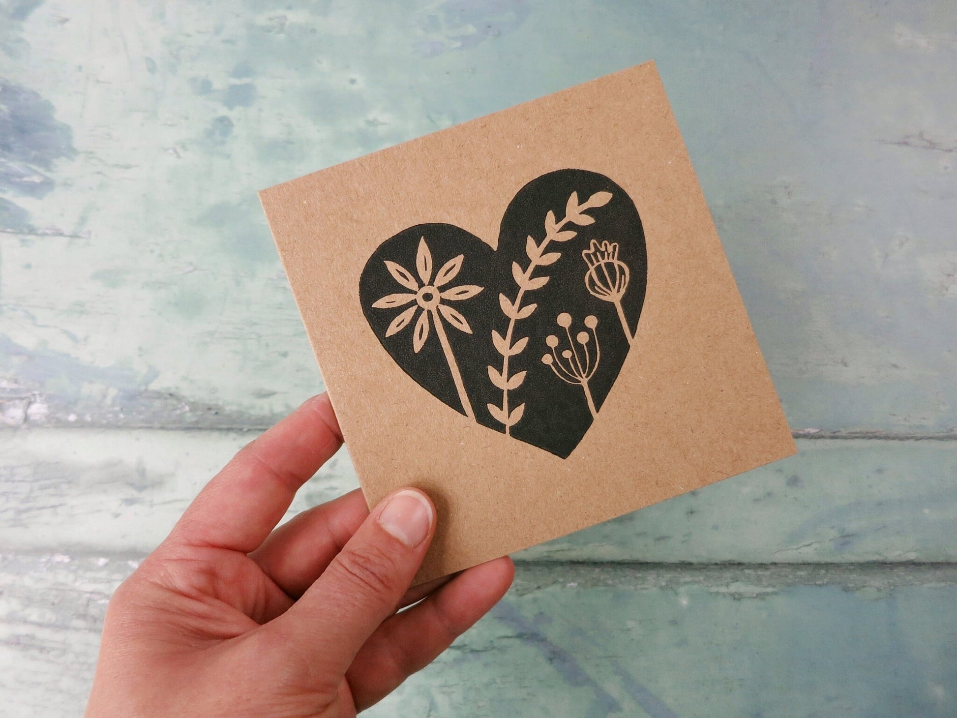Hand holding Kraft paper card with an original black lino printed heart design featuring floral elements against a textured surface.