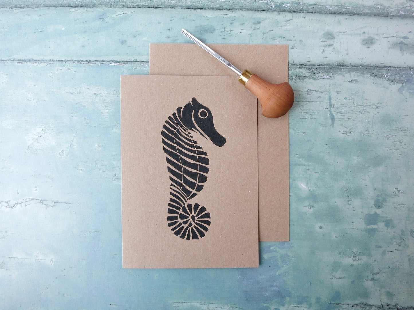 Kraft paper card with an original seahorse lino print laid on a textured blue surface. Photo includes lino cutting tool