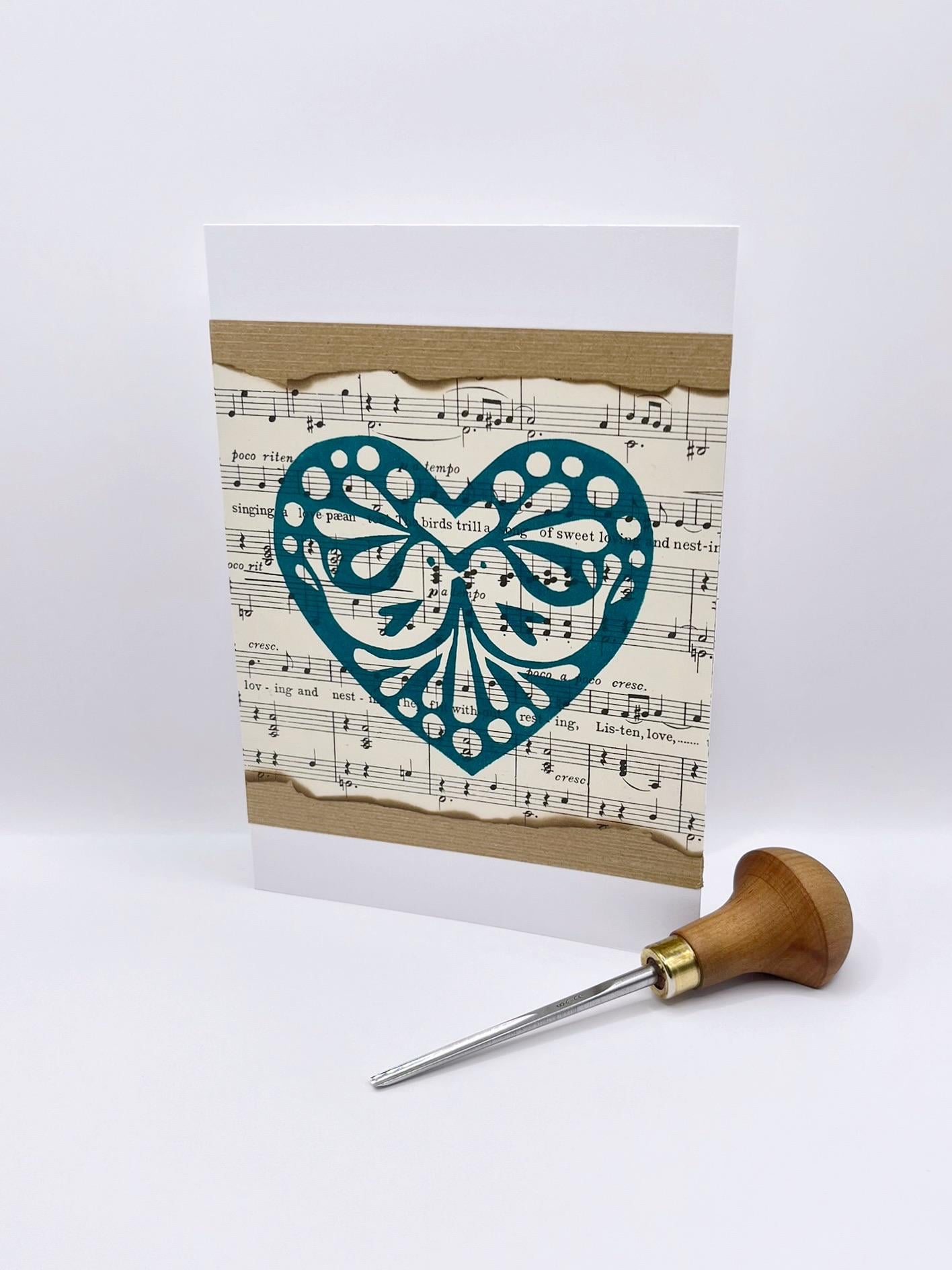Card with decorative heart design on sheet music. Photo includes lino cutting tool laid on a white background.