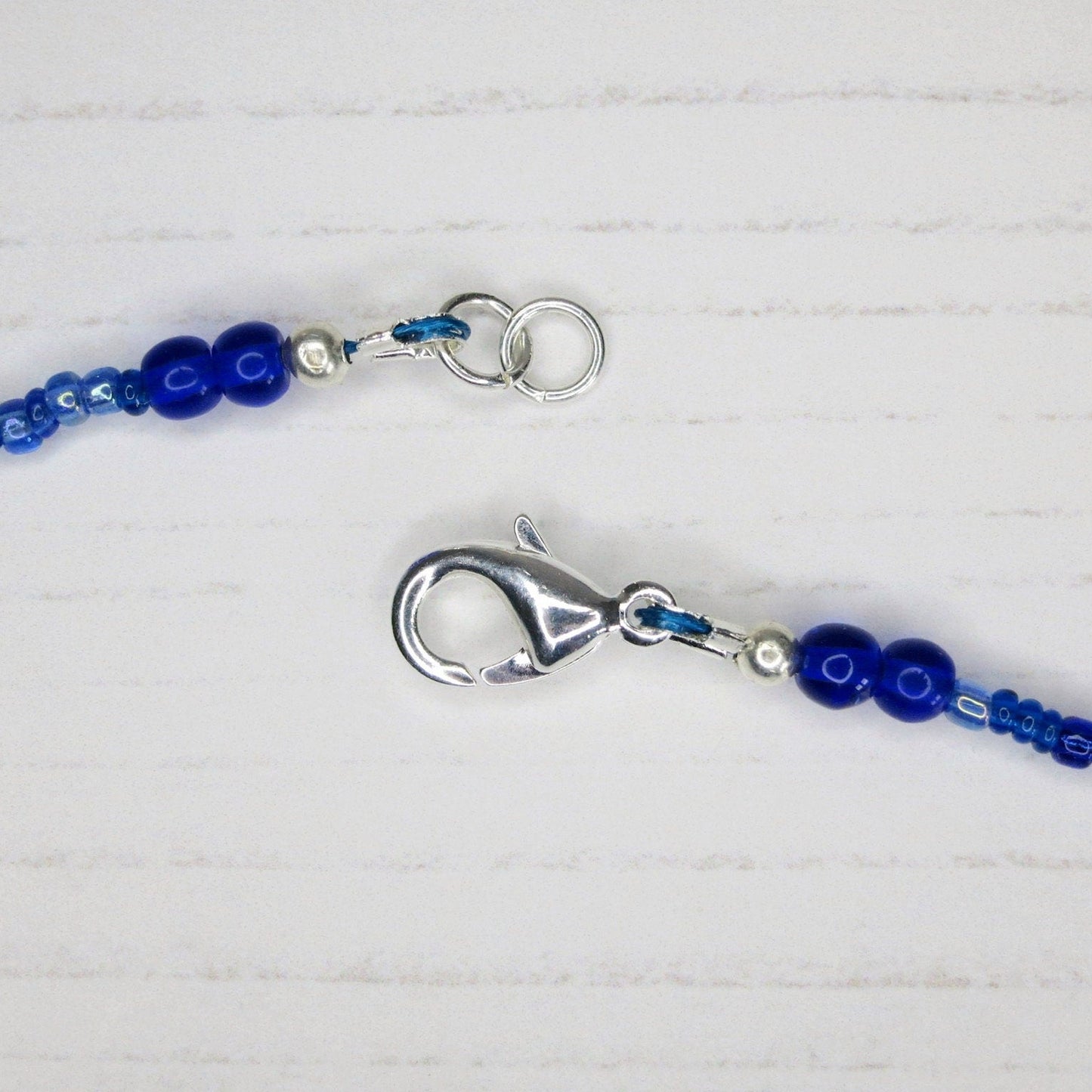 Close up of the silver plated catch on the blue seed bead necklace laid on a pale background.