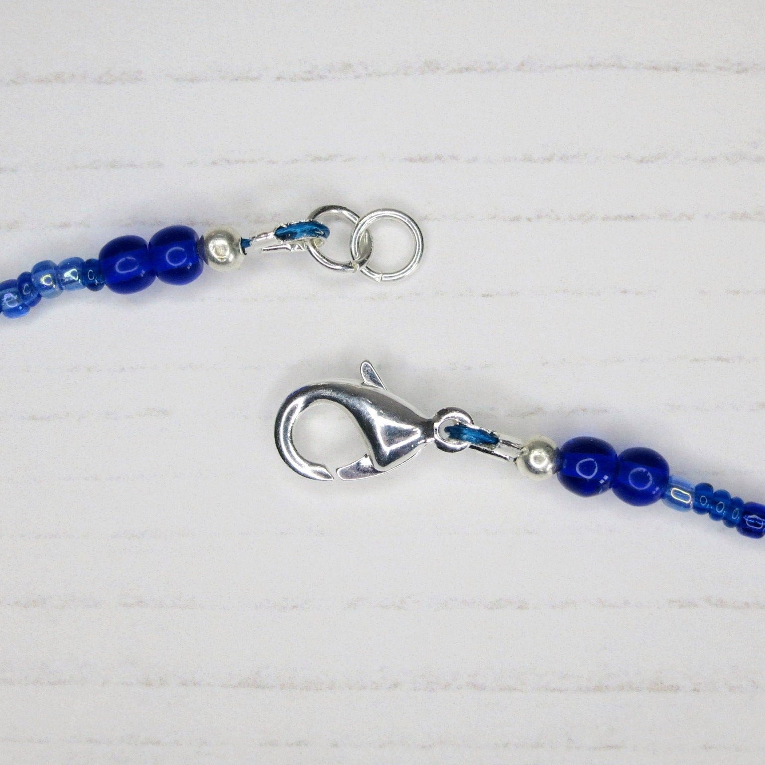 Close up of the silver plated catch on the blue seed bead necklace laid on a pale background.
