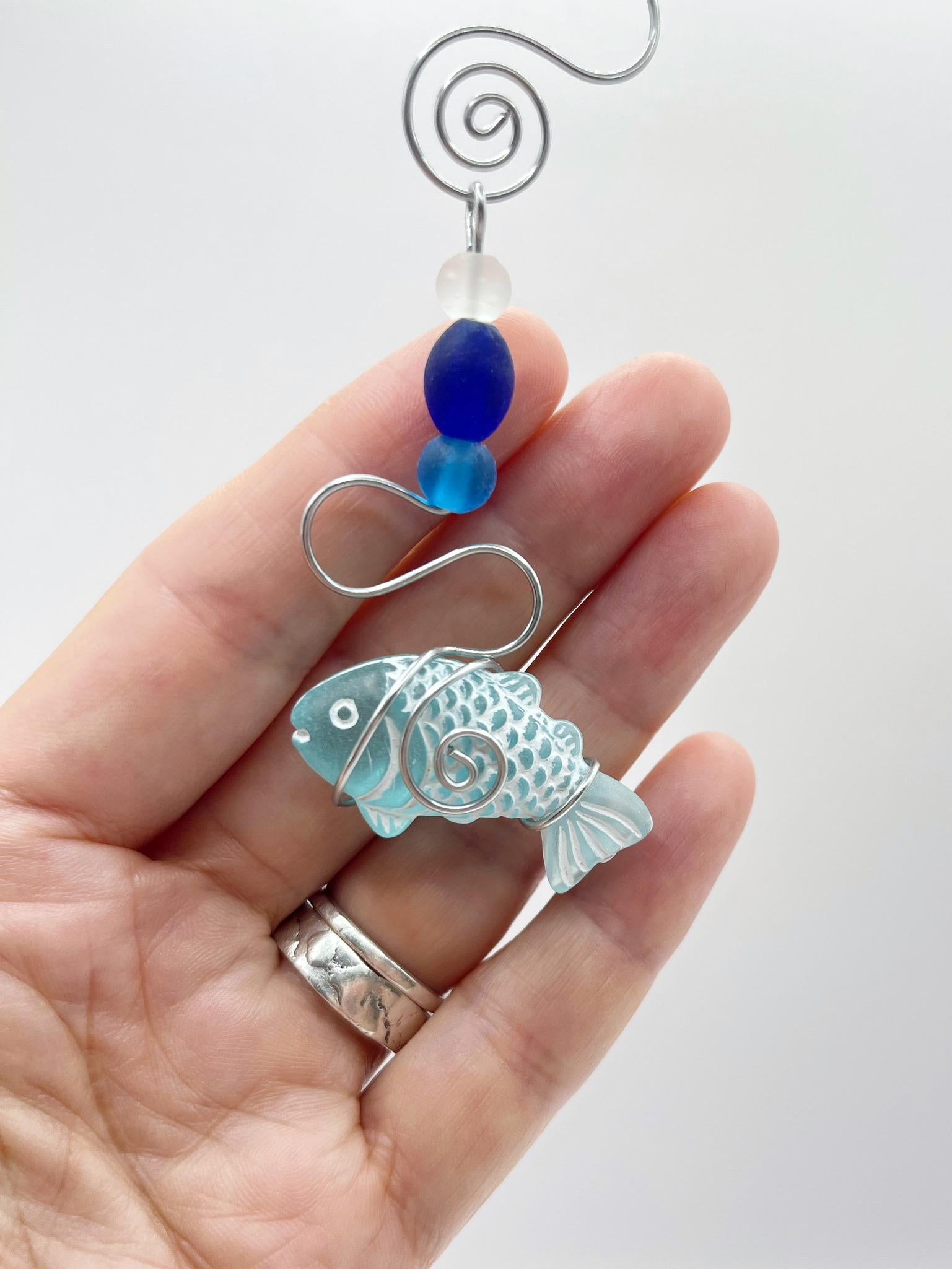 Decorative hanging fish ornament with wire and blue beads held in a hand against a white background