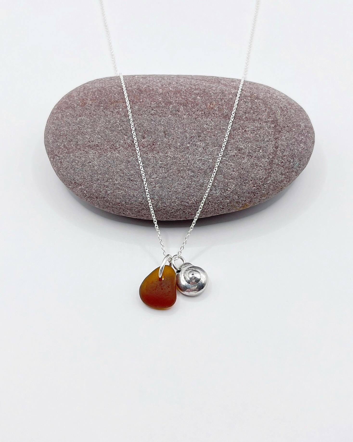 Necklace with burnt orange sea glass and a silver shell pendant, laid infant of a purple pebble on a white surface