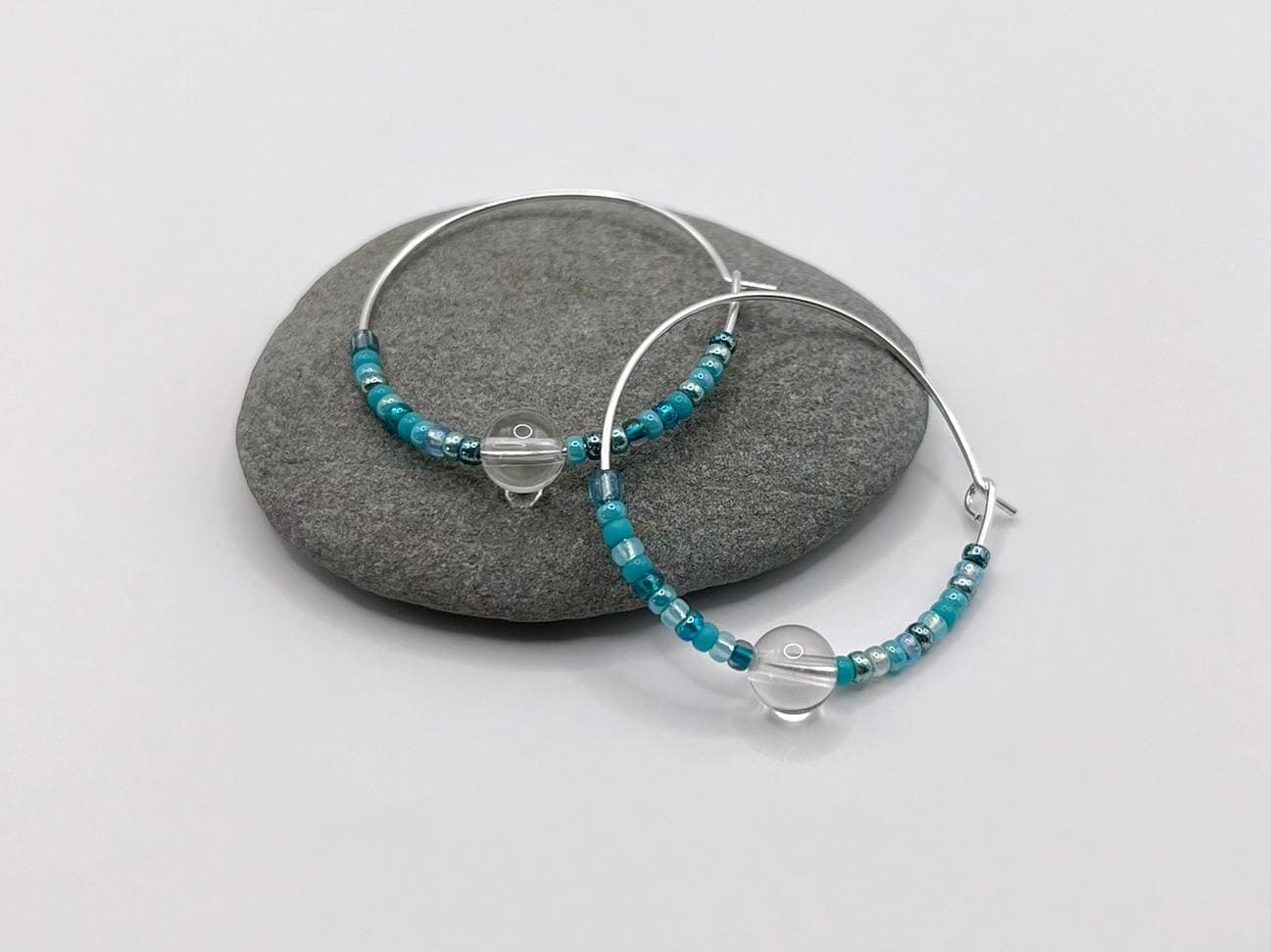 Silver hoop earrings with turquoise seed beads and crystal laid on a grey pebble against a white background