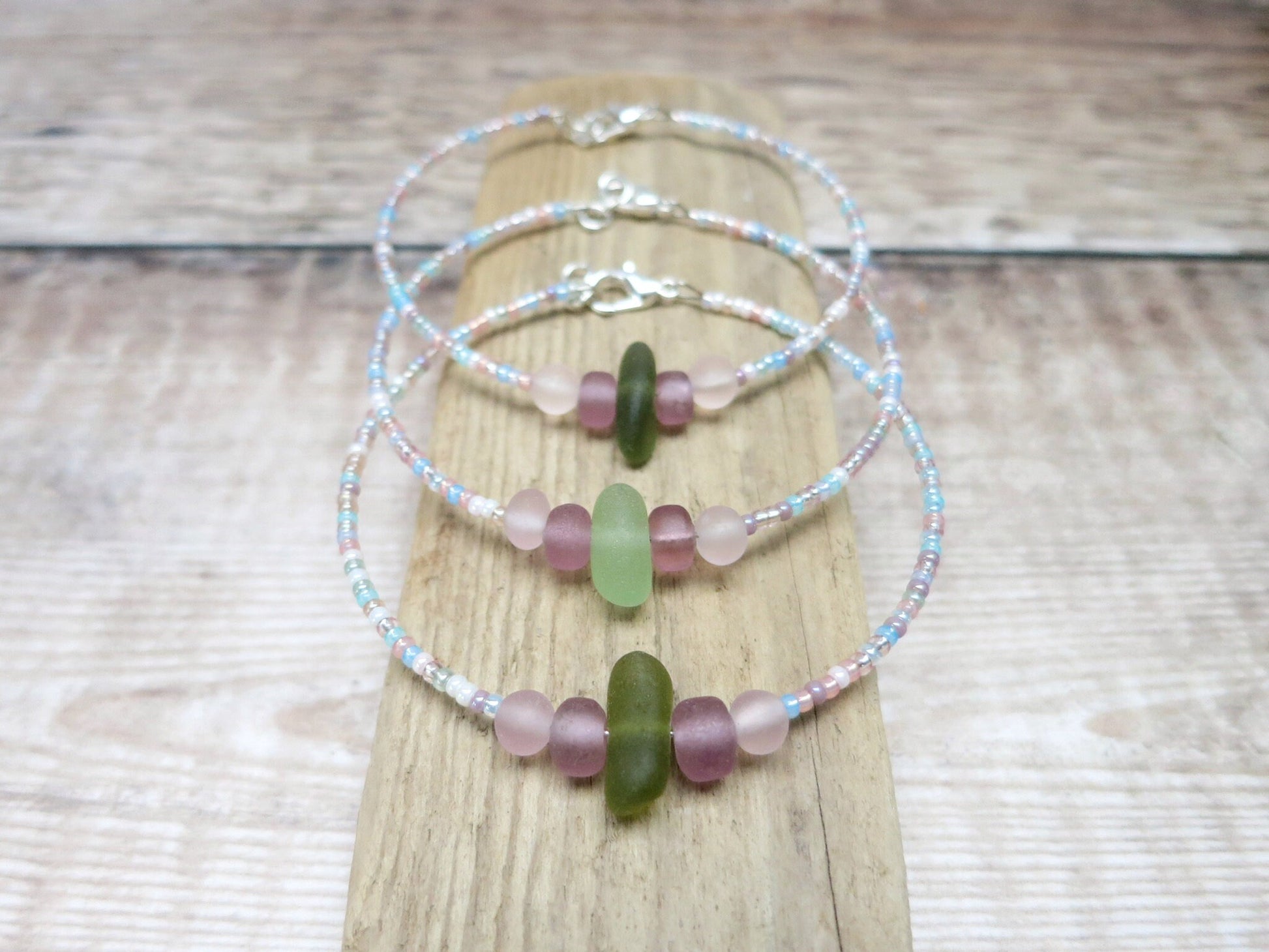 Three pastel seed beaded bracelets with green sea glass laid on a driftwood 