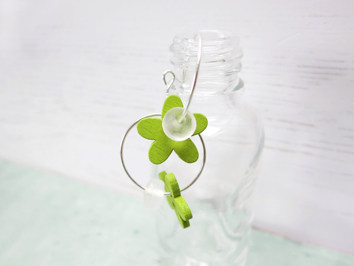 Close up of silver hoop earrings with green wooden flowers and a glass bead hung on a glass bottle