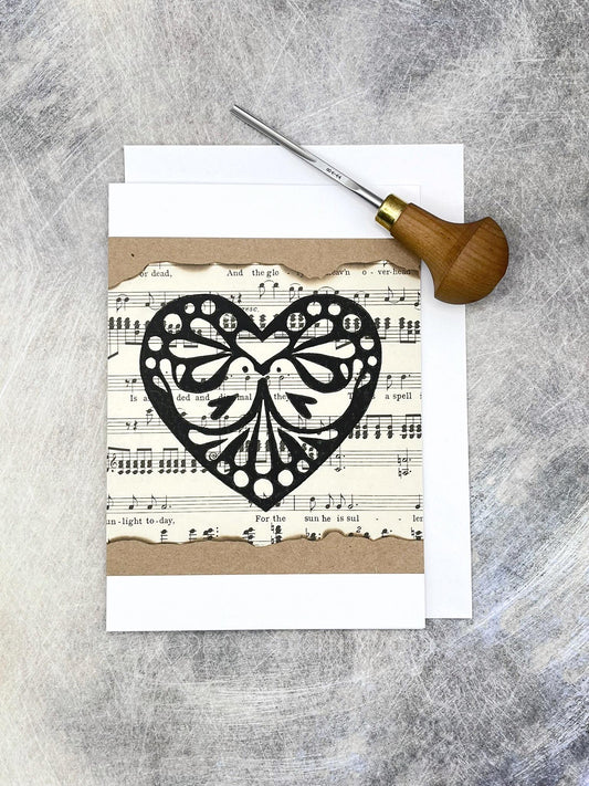 Card with original lino printed heart design on sheet music. Photo includes lino cutting tool laid on textured background.