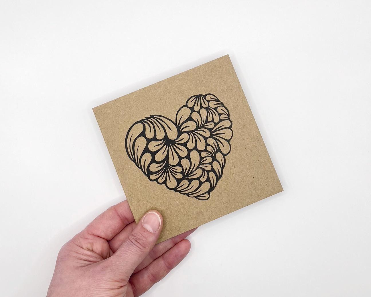Hand holding a Kraft card with a black heart design against a white background