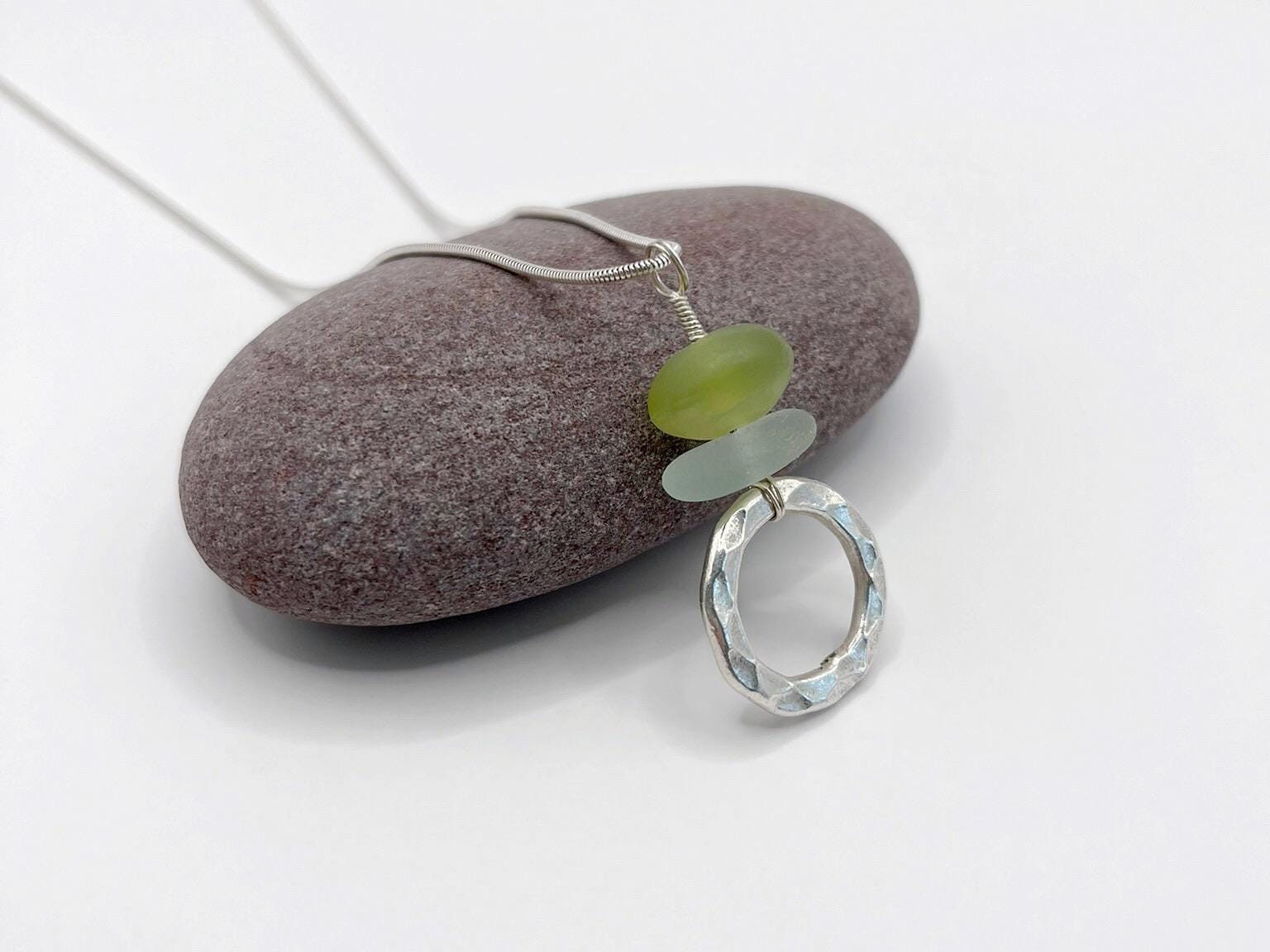 Necklace with a silver ring pendant, jade bead and sea glass laid against a purple pebble on a white background
