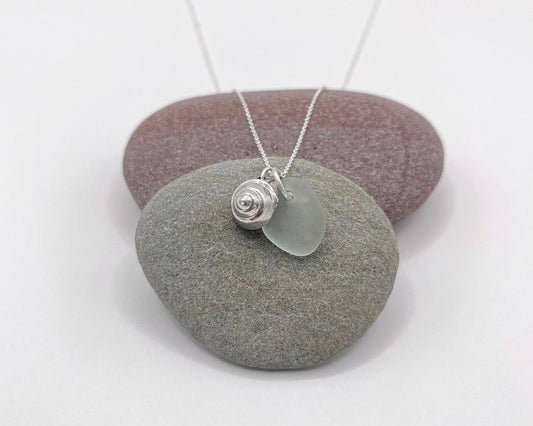 Necklace with a silver shell and seaglass pendant laid on a pebble against a white background