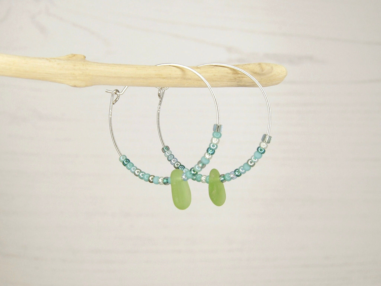 Pair of hoop earrings with green sea glass and turquoise seed beads hanging on driftwood against a pale background.