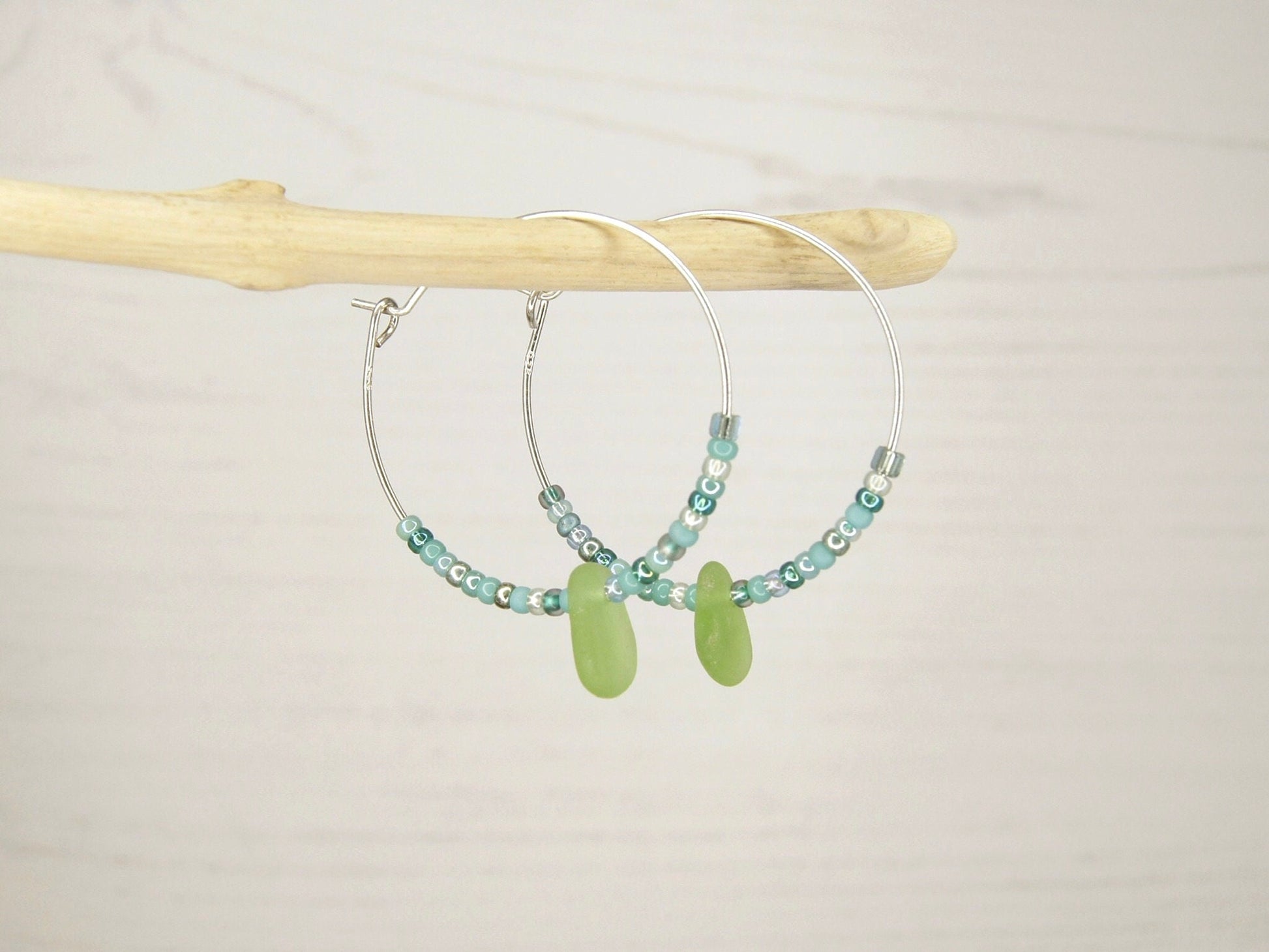 Pair of hoop earrings with green sea glass and turquoise seed beads hanging on driftwood against a pale background.