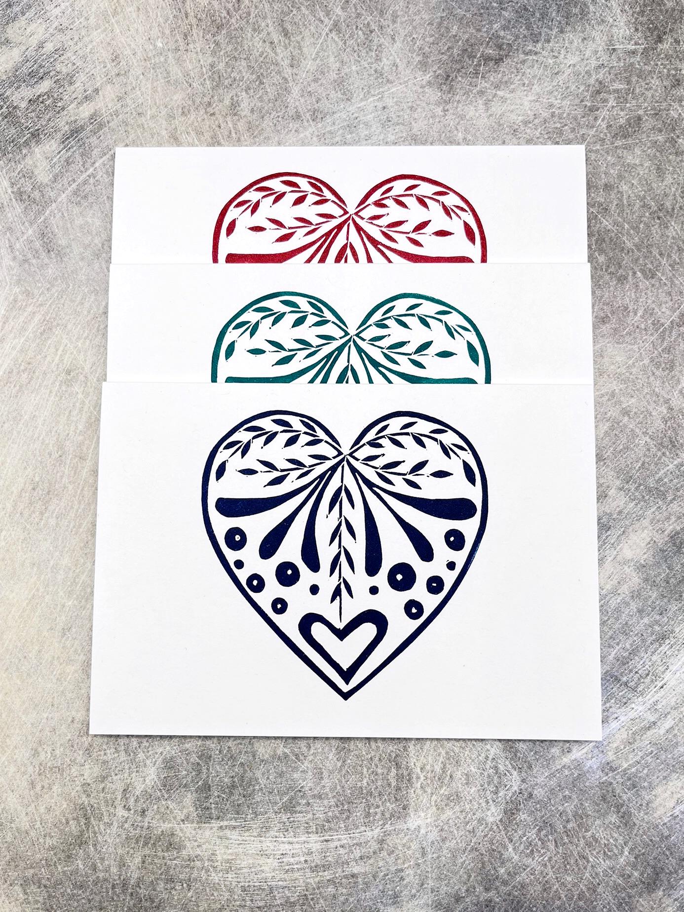 Set of three white cards with original lino printed heart designs in blue, green and red. Laid on a textured surface