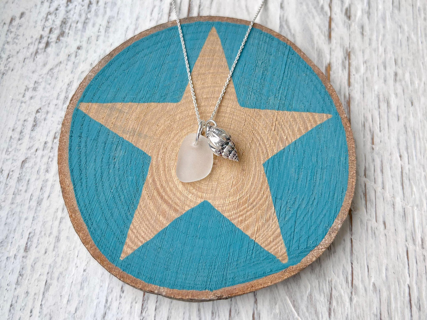 Necklace with white sea glass and silver shell pendant laid on a star printed slice of wood against a textured surface