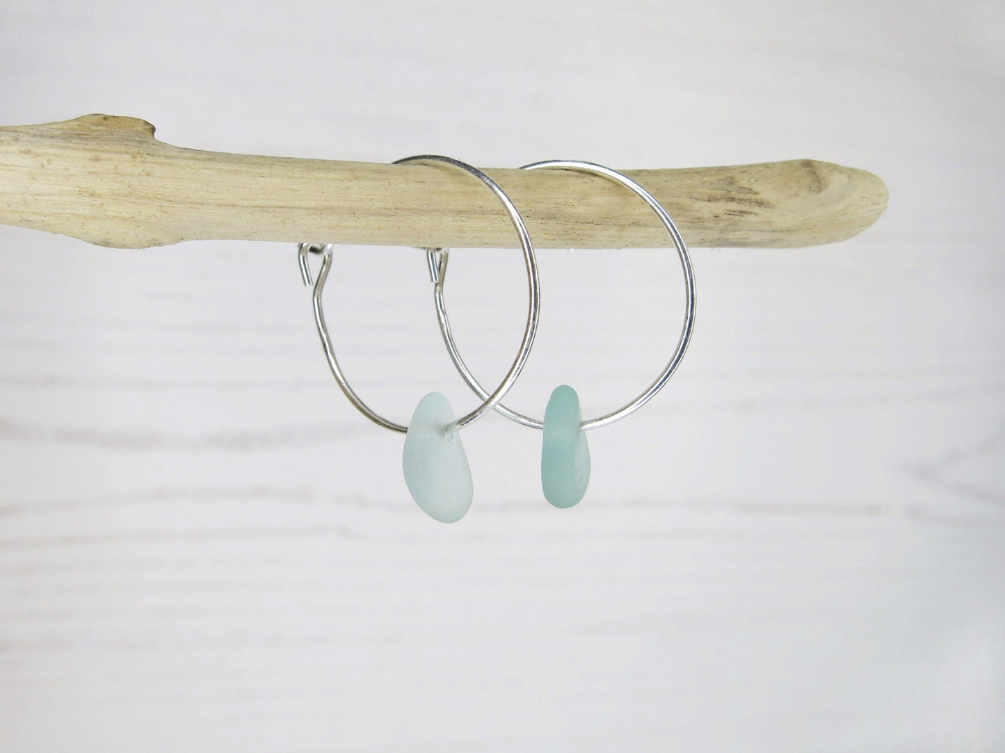 Silver hoop earrings with sea foam sea glass hanging on driftwood against a white background