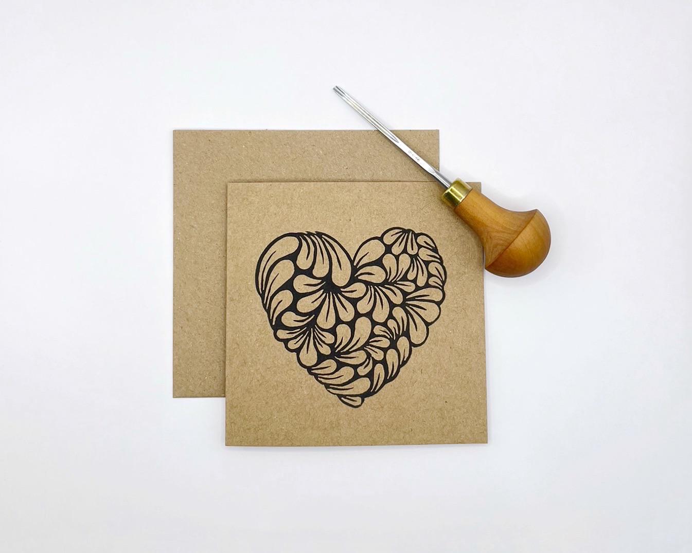 Kraft paper card with original lino printed  heart laid on a white background. Photo includes lino cutting tool used.
