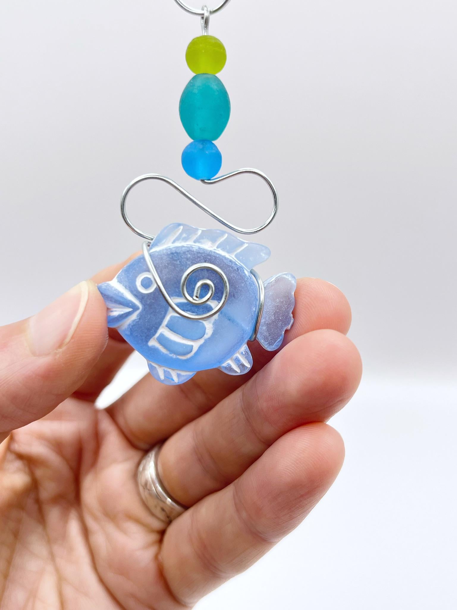 Decorative hanging ornament with blue fish, wire and colourful beads held in a hand against a white background