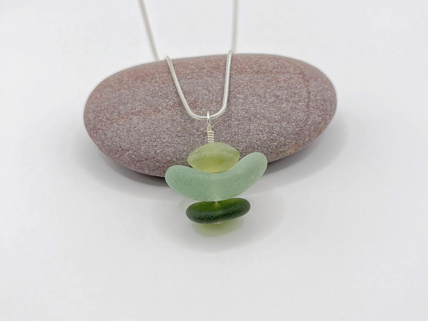 Necklace with green sea glass and Jade pendant sat against a purple pebble on a white background