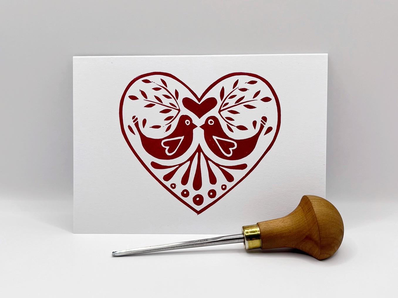 White card with an original lino printedred heart design featuring two birds stood against a white background. Photo includes lino cutting tool 