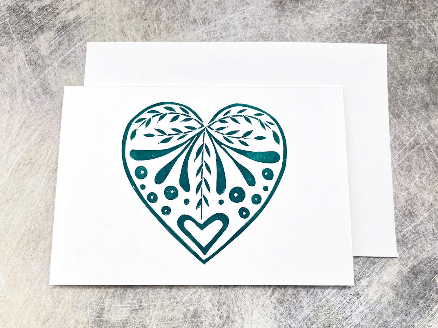 White card with an original lino printed green heart design laid on a textured surface. 
