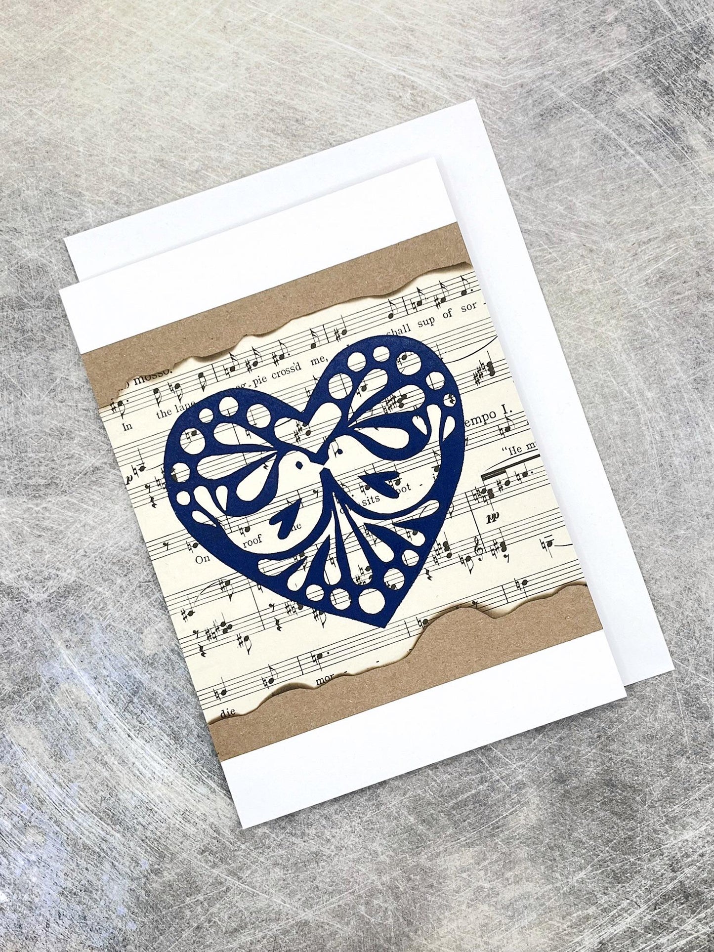 Card with blue original lino printed heart design on sheet music background, laid on a grey textured background.