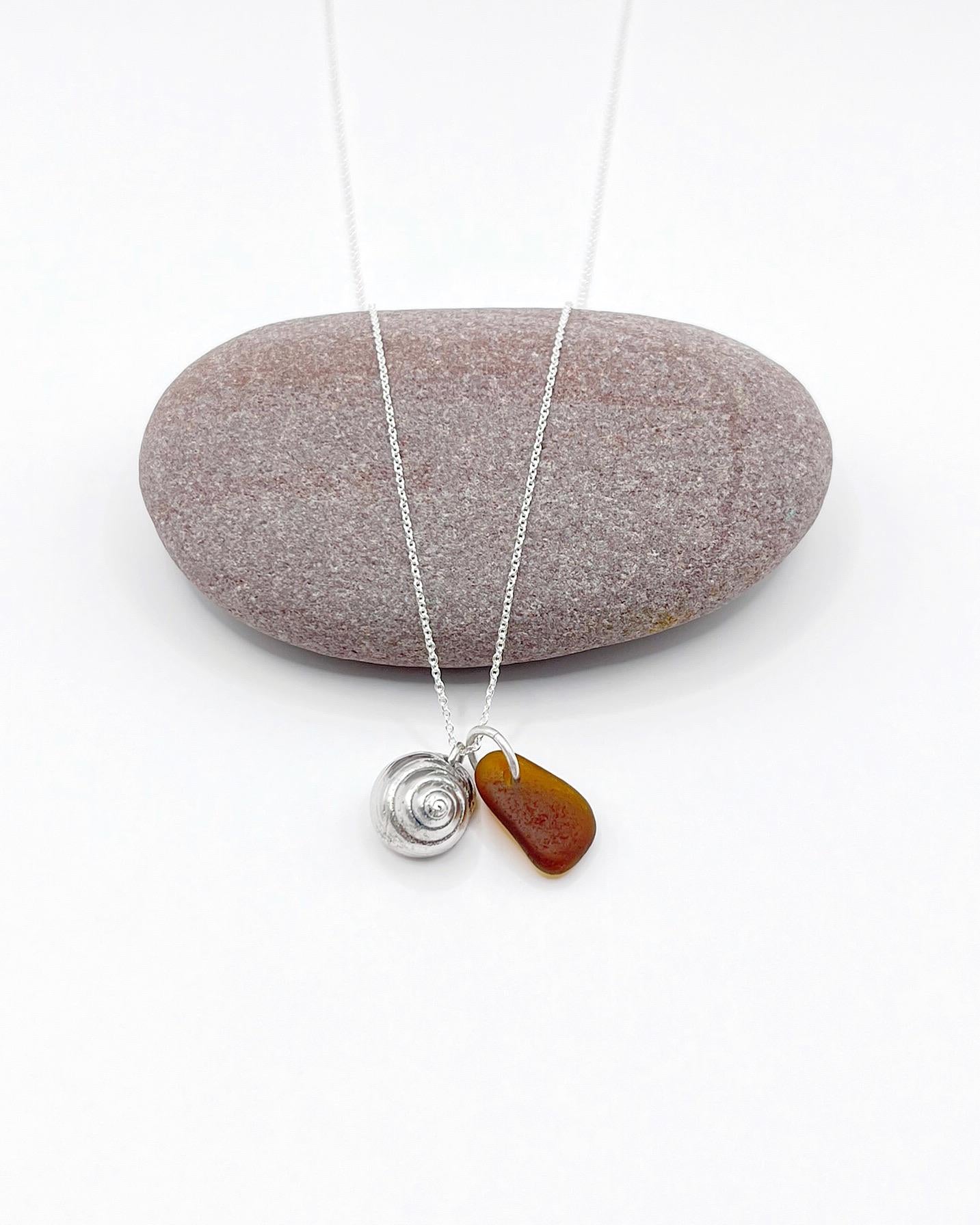 Silver necklace with burnt orange sea glass and a silver shell pendant laid in front of a purple pebble on a white background