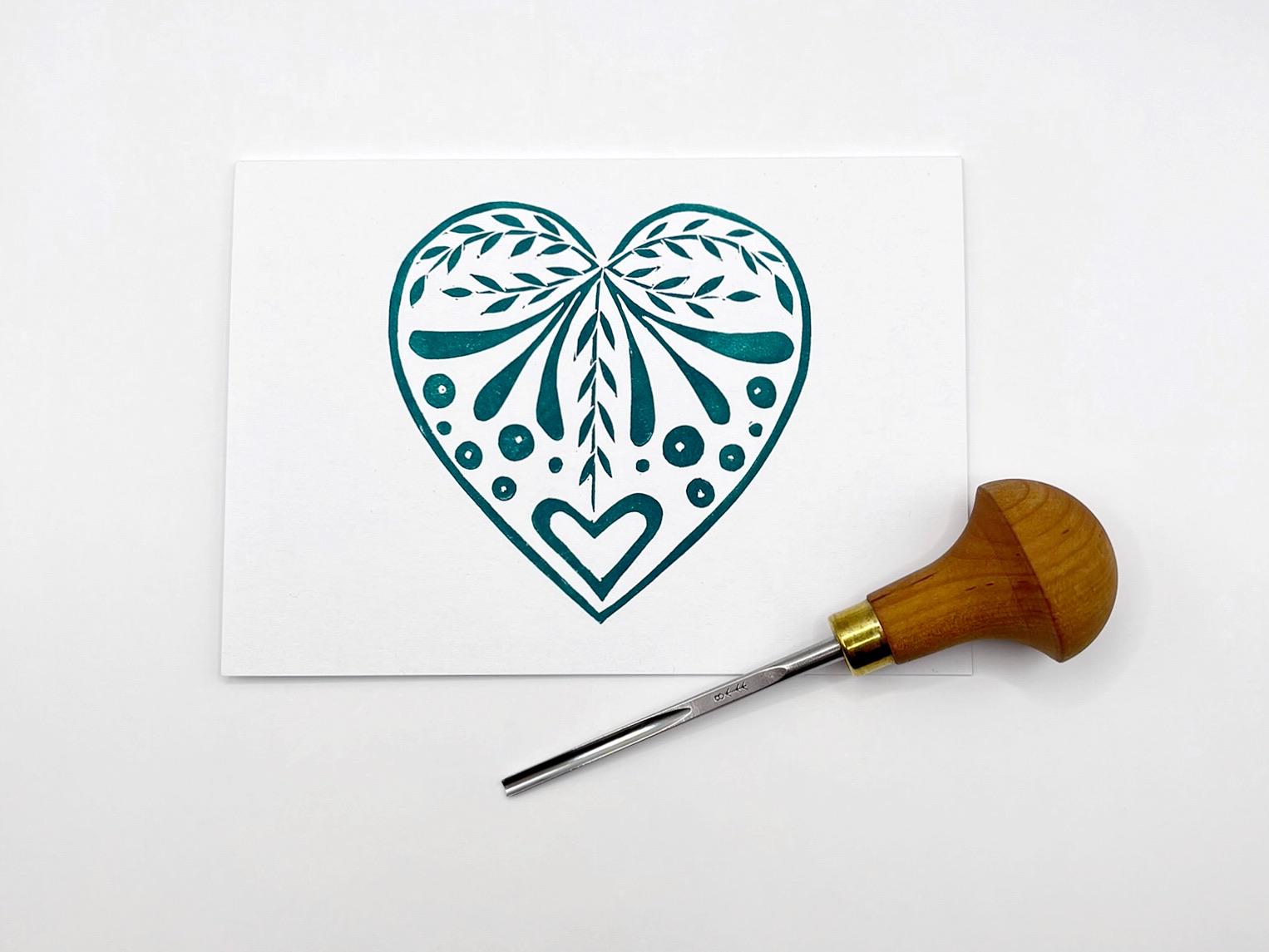 White card with an original lino printed green heart design laid on a white surface. Photo includes lino cutting tool.