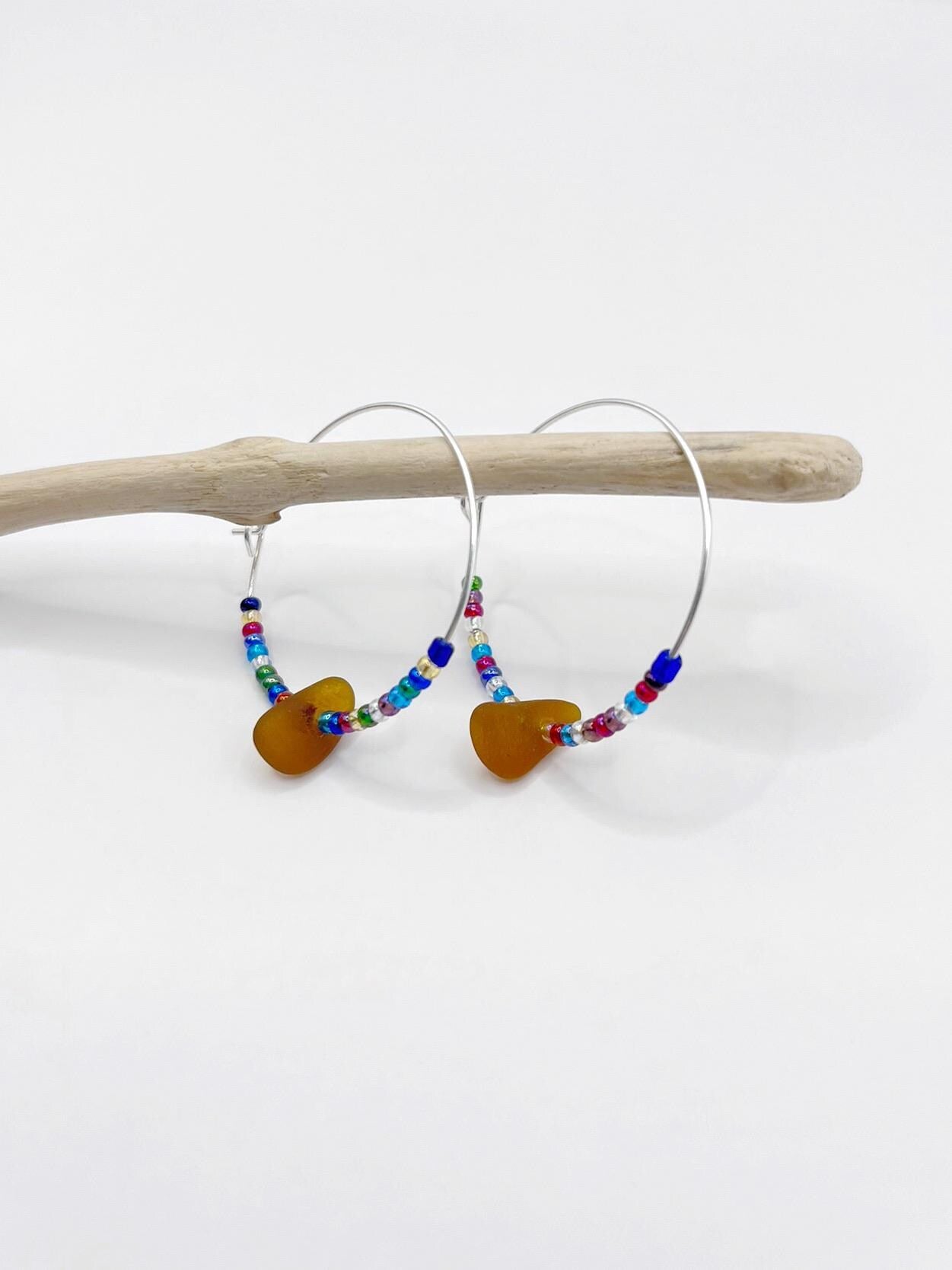 Rainbow seed bead and burnt orange sea glass hoop earrings hanging on driftwood against a white background