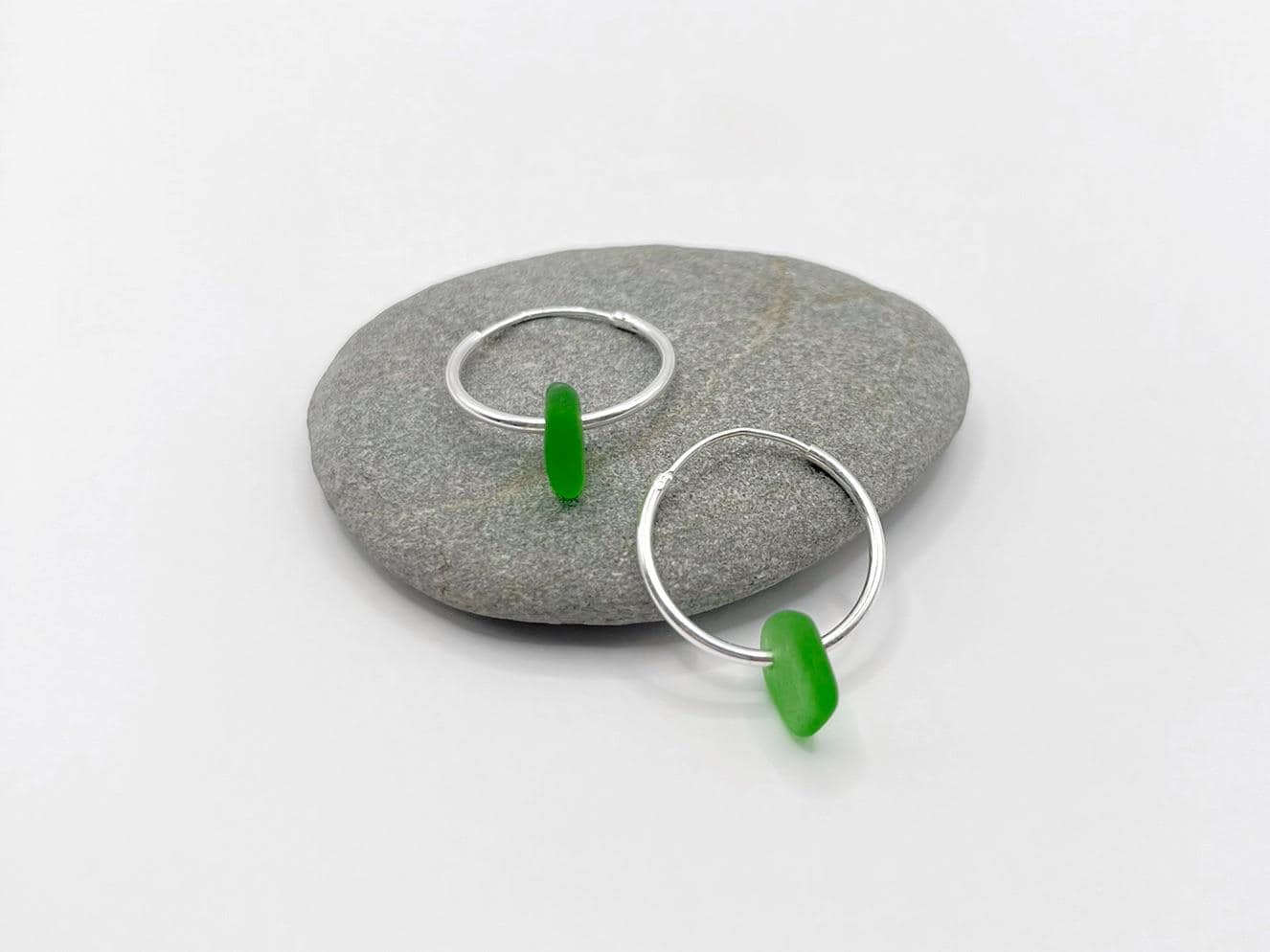 Silver hoop earrings with green sea glass laid on a grey pebble against a white background