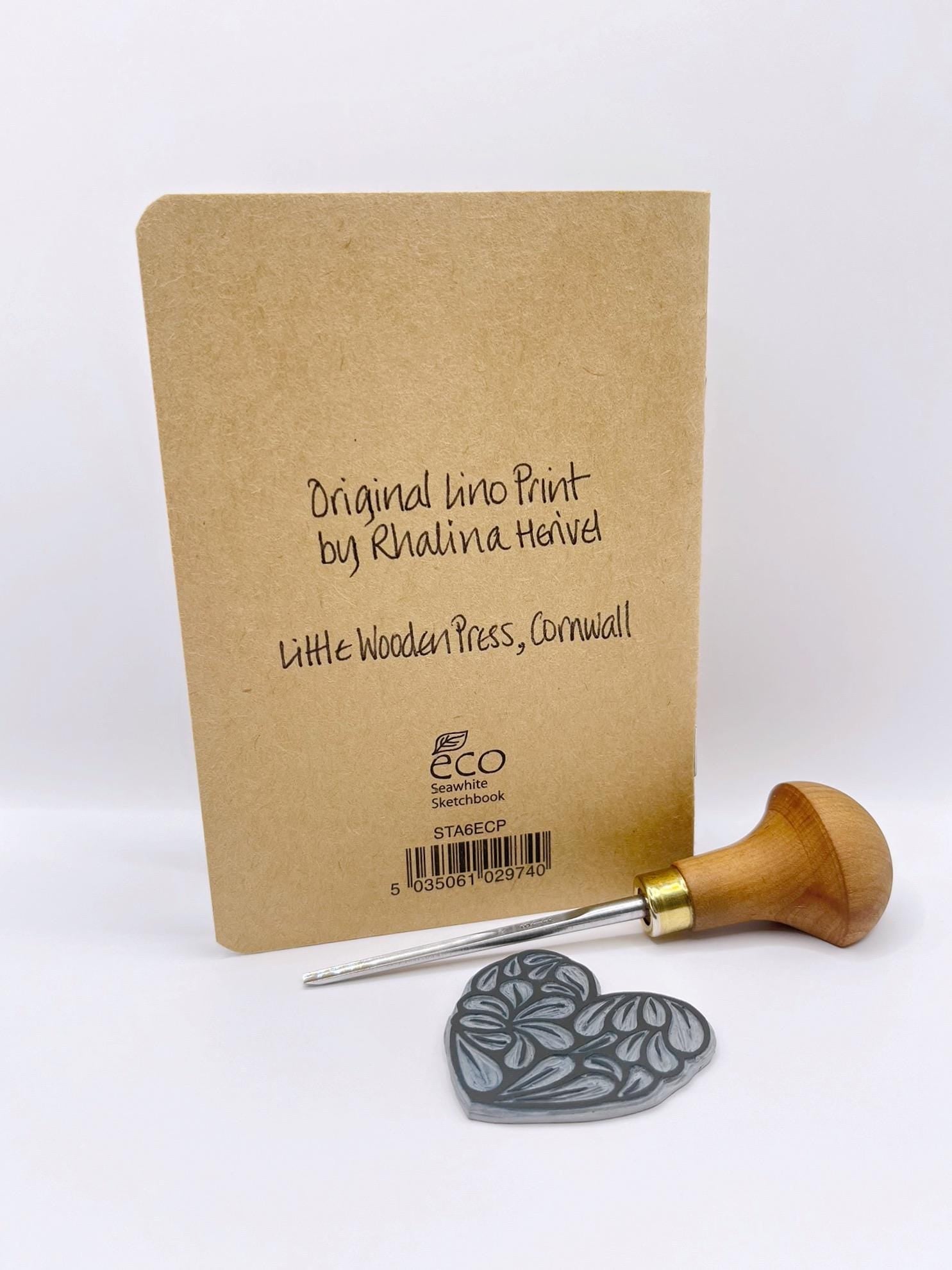 Showing the back of the Kraft notebook with 'Original Lino Print by Rhalina Hevel' text, a wooden tool, and a gray stamp on a white background.
