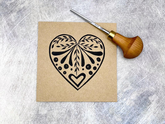 Card with an original lino printed black heart design on Kraft card laid on a textured surface. Photo includes lino cutting tool used.