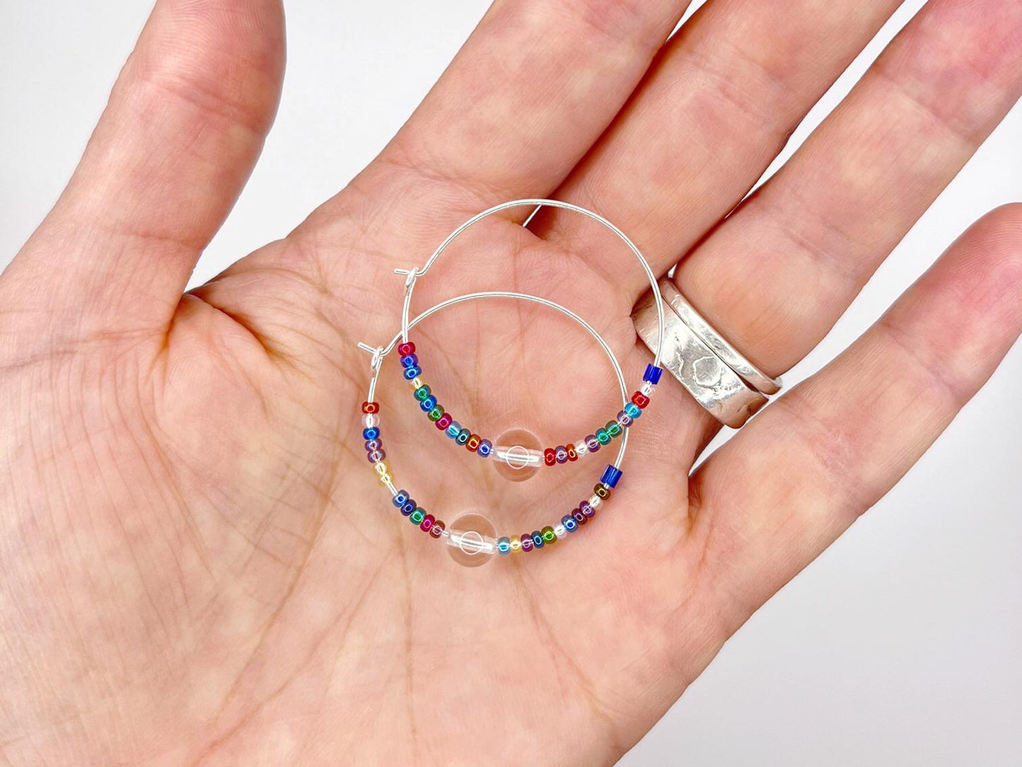 Hand holding a pair of hoop earrings with colourful beads on a plain background
