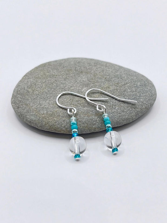 Pair of earrings with turquoise and clear crystal beads laid on a grey pebble.